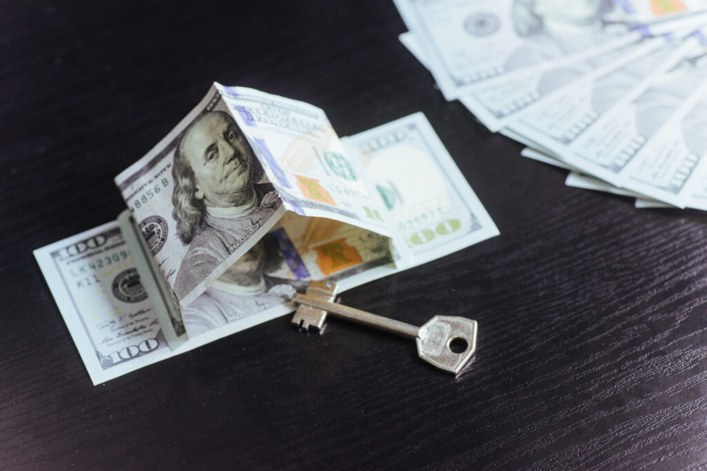 A small house made from folded $100 bills with a key placed in front, symbolizing the financial aspects of homeownership, hidden costs, and real estate investments.